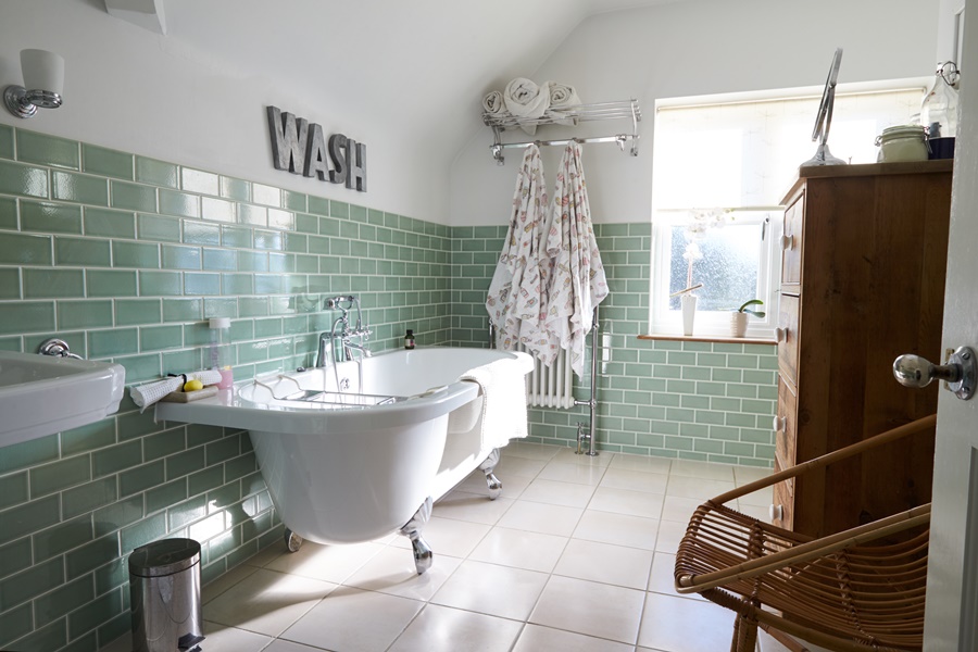 Azulejos tipo metro en verde menta, bañera exenta con grifería cromada y muebles de madera. Todo muy alineado con lo descrito en los apartados sobre revestimientos y mobiliario.