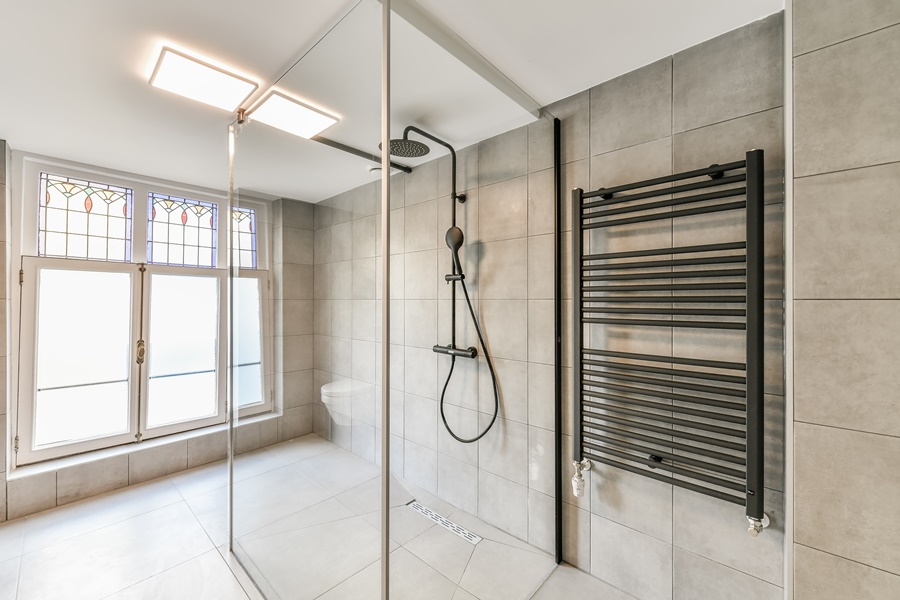Columna de ducha con rociador y teleducha integrada en un baño amplio con mampara de cristal, azulejos grises y ventana con vidrieras decorativas.